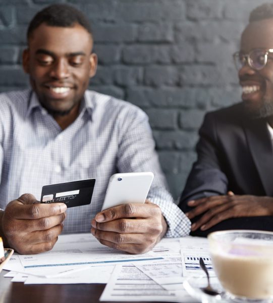 Successful happy African-American businessman paying bill with credit card online via internet, making transaction, using mobile bank application on cell phone during meeting with his partner at cafe